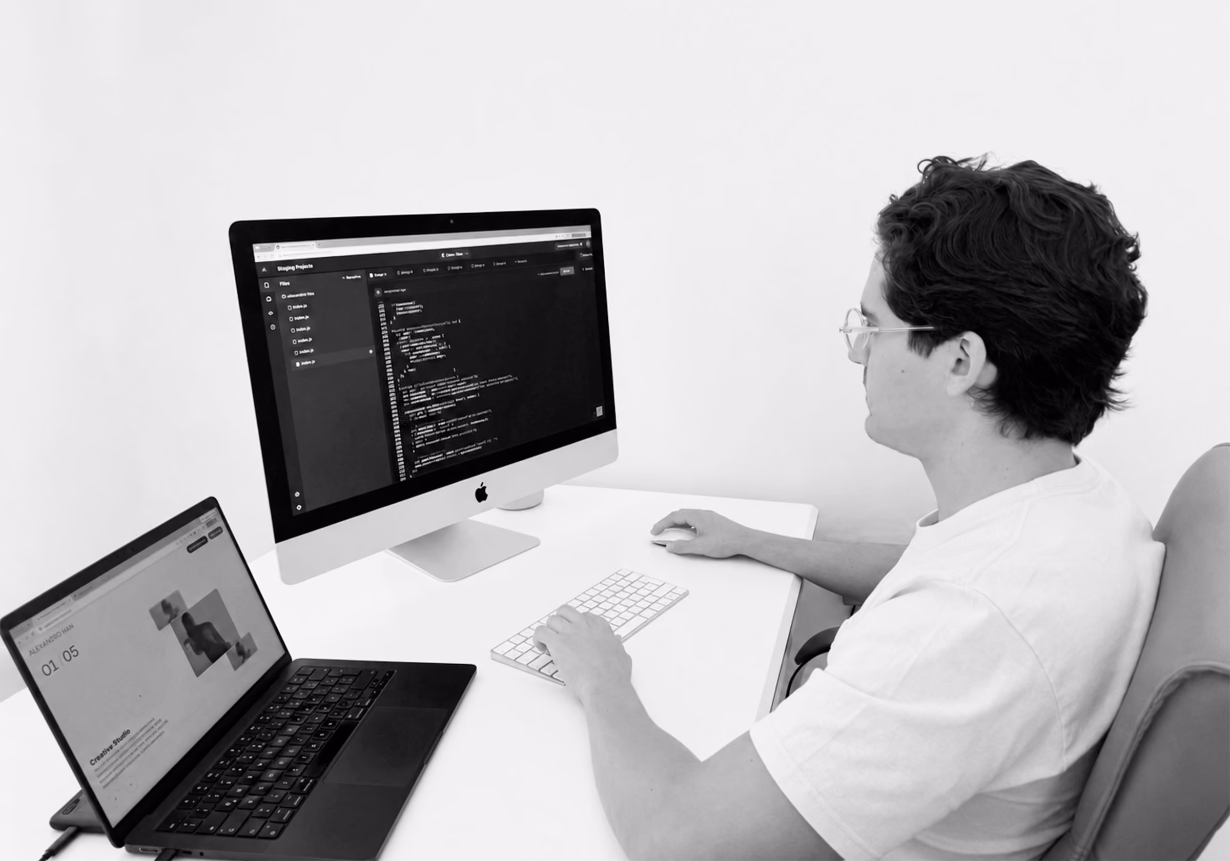 Man at a white desk with code on a desktop monitor and a laptop beside it.